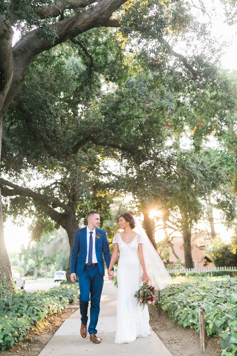 May include: A bride and groom walking hand-in-hand down a path lined with greenery. The bride is wearing a white lace dress with a veil and carrying a bouquet of red flowers. The groom is wearing a blue suit with a blue tie. The couple is smiling and looking at each other.