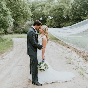 May include: A bride and groom stand on a dirt path, the bride's veil billowing in the wind behind them. The groom is wearing a dark suit and the bride is wearing a white wedding dress with a bouquet of white flowers.