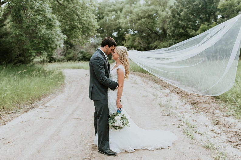 May include: A bride and groom stand on a dirt path, the bride's veil billowing in the wind behind them. The groom is wearing a dark suit and the bride is wearing a white wedding dress with a bouquet of white flowers.