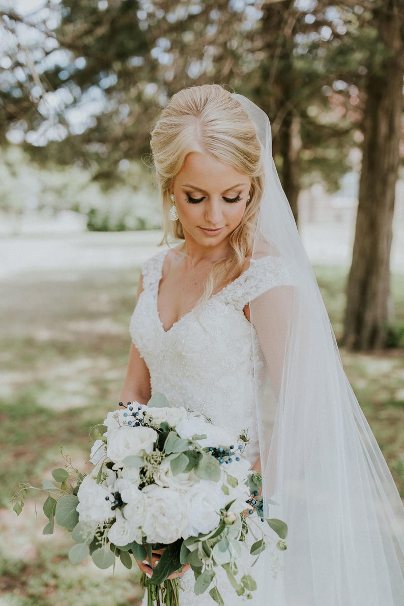 May include: A bride in a white lace wedding dress with a long veil holds a bouquet of white flowers and greenery.