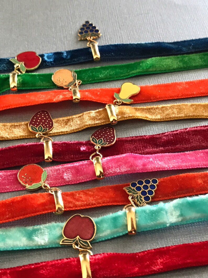 May include: A set of ten velvet ribbon chokers with gold clasps and enamel fruit charms. The charms include a bunch of grapes, a pear, a strawberry, an apple, and an orange.