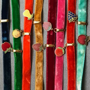 Op de afbeelding: Tien fluwelen lint-kettingen met gouden bedels met verschillende vruchten, waaronder aardbeien, druiven, appels, peren en sinaasappels. De linten zijn in verschillende kleuren, waaronder rood, groen, blauw, turquoise en roze.