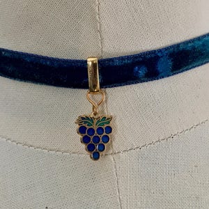 Op de afbeelding: Een blauwe fluwelen chokerketting met een gouden bedel in de vorm van een tros druiven. De druiven zijn blauw en groen.