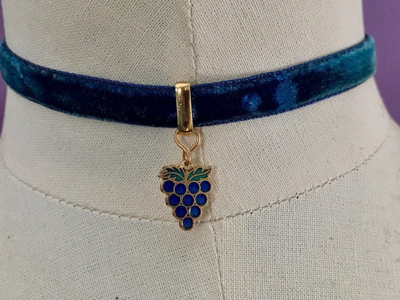 Op de afbeelding: Een blauwe fluwelen chokerketting met een gouden bedel in de vorm van een tros druiven. De druiven zijn blauw en groen.