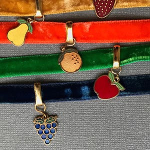 Op de afbeelding: Vijf fluwelen lint-chokers met verschillende gekleurde bedels. De bedels zijn een peer, een sinaasappel, een appel en een tros druiven. De chokers zijn in de kleuren goud, oranje, groen, blauw en zwart.