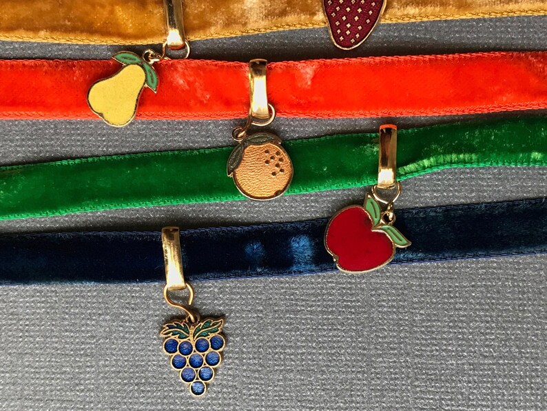 Op de afbeelding: Vijf fluwelen lint-chokers met verschillende gekleurde bedels. De bedels zijn een peer, een sinaasappel, een appel en een tros druiven. De chokers zijn in de kleuren goud, oranje, groen, blauw en zwart.