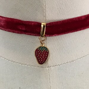 Op de afbeelding: Een rode fluwelen choker met een gouden bedel in de vorm van een rode aardbei met groene bladeren.