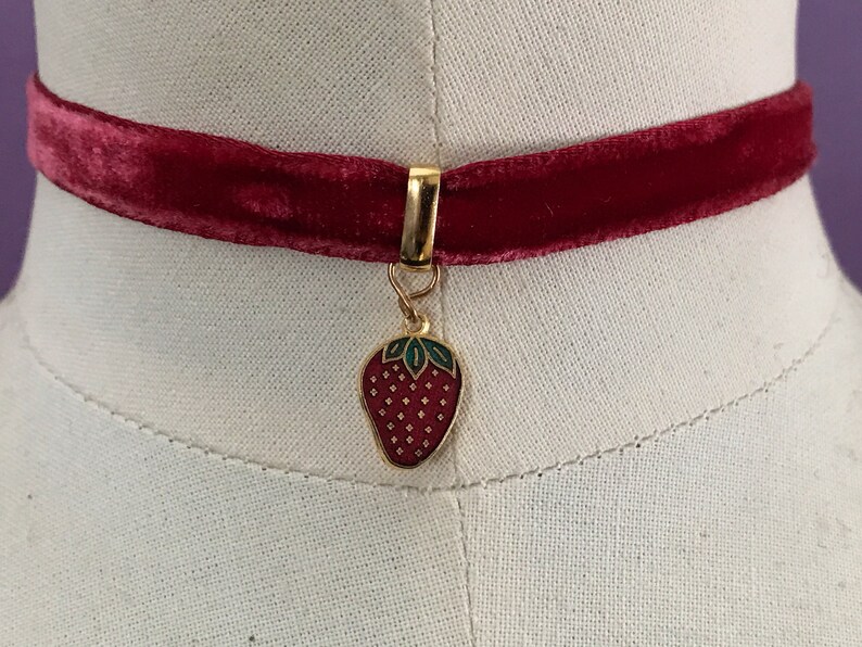 Op de afbeelding: Een rode fluwelen choker met een gouden bedel in de vorm van een rode aardbei met groene bladeren.