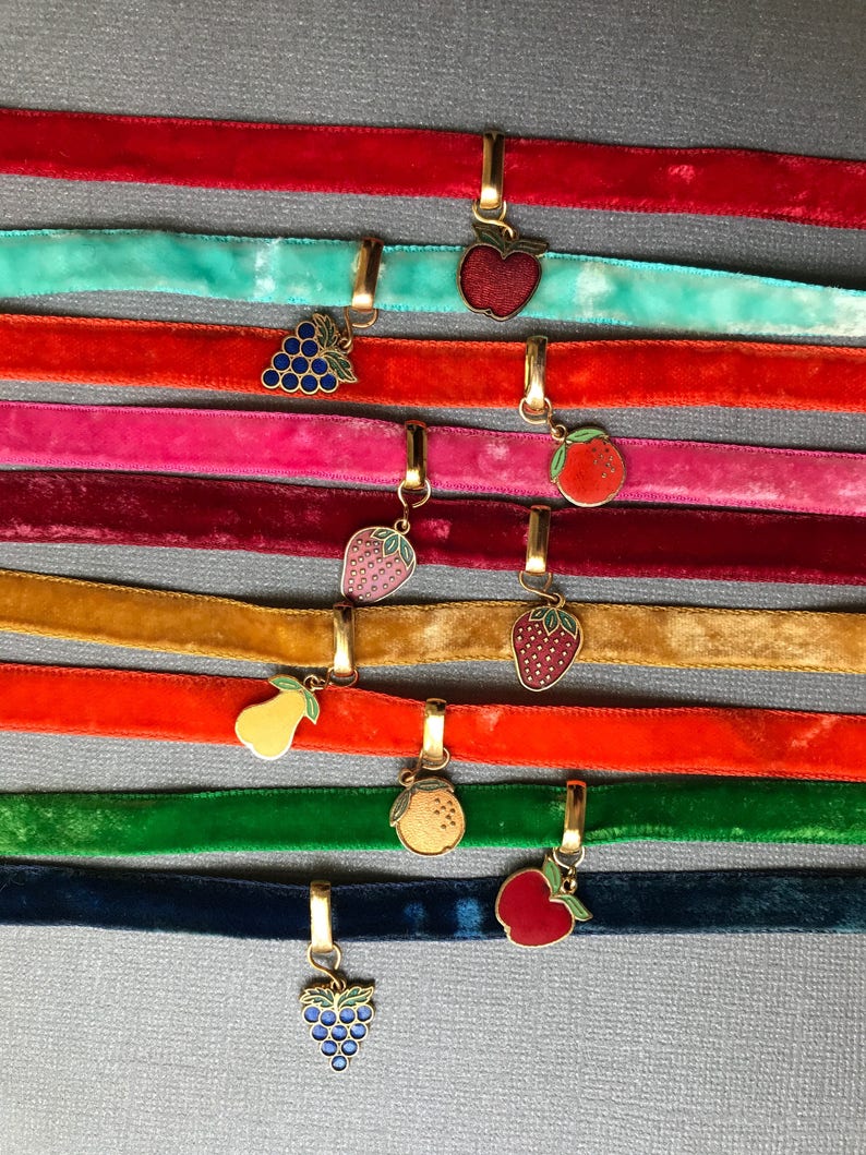 May include: Ten velvet ribbon chokers with gold charms featuring different fruits and berries. The charms include an apple, grapes, a strawberry, a pear, an orange, and a lemon.