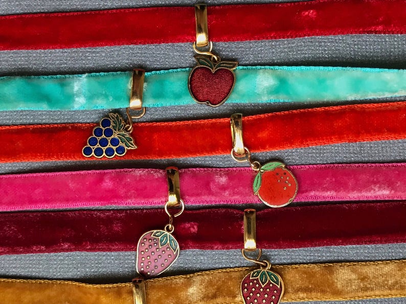 Op de afbeelding: Zes fluwelen lint-choker met gouden sluitingen en ge&euml;mailleerde fruitbedels. De bedels omvatten een appel, druiven, een sinaasappel en twee aardbeien.
