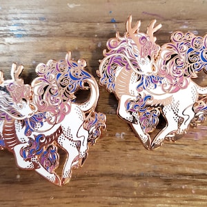 May include: Two gold-tone enamel pins featuring a white mythical creature with purple, blue, and pink swirling designs. The creature has a long, flowing mane and tail, and a single antler on its head.