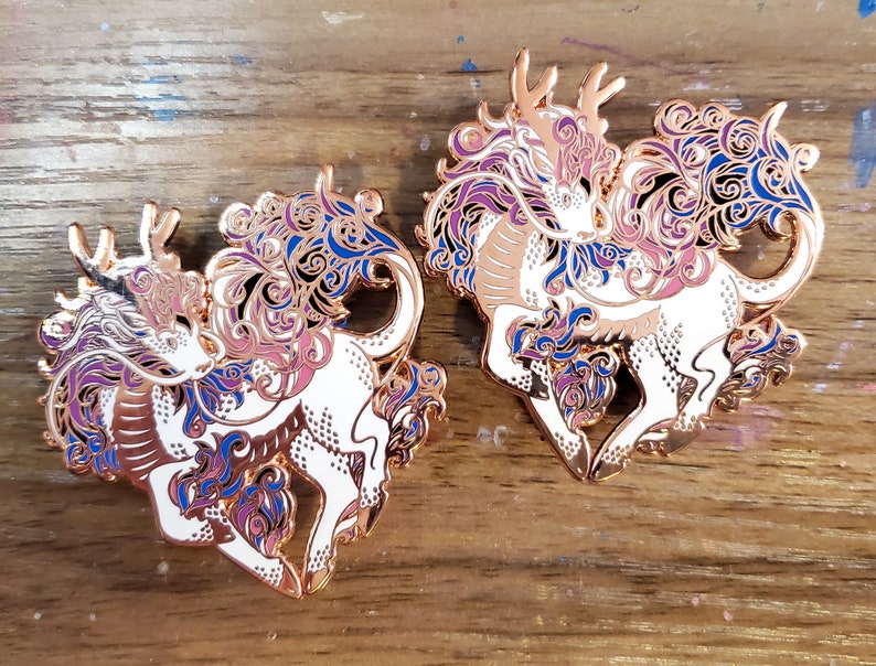 May include: Two gold-tone enamel pins featuring a white mythical creature with purple, blue, and pink swirling designs. The creature has a long, flowing mane and tail, and a single antler on its head.