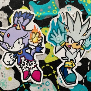 May include: Two cartoon character stickers, one of a purple and blue cat with a fiery orange tail and the other of a silver hedgehog with blue flames. Both characters are in a fighting pose.