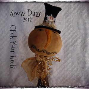 May include: A handcrafted snowman figure with a tan head, black top hat, and gold scarf. The snowman has painted facial features and is embellished with gold tinsel. The text "Snow Daze 2012" and "Click Your Heels" are visible.