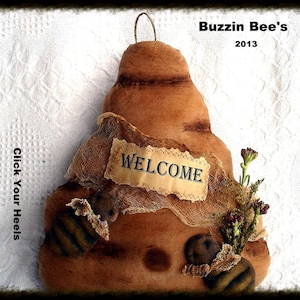Puede incluir: Un adorno de pared con forma de colmena de tela marrón con un letrero de bienvenida. El letrero dice "WELCOME". La colmena tiene un aspecto rústico y texturizado, con elementos decorativos como tela y flores secas. También es visible el texto "Buzzin Bee's 2013".