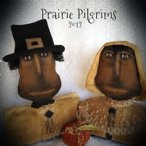 May include: Two handmade rag dolls dressed as pilgrims. The male doll wears a black hat and the female doll wears a brown hooded cloak. Both dolls have painted faces and are holding apples.
