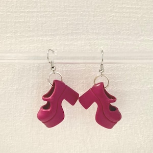May include: A pair of magenta platform shoe earrings. Each earring features a miniature, high-heeled shoe with a chunky heel and a Mary Jane strap. The earrings are suspended from silver-colored fish hook ear wires.