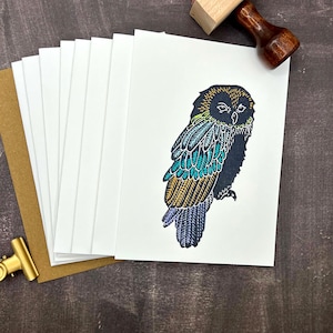 May include: A stack of white greeting cards featuring a stylized owl illustration with intricate feather details in shades of yellow, teal, and purple. The cards are accompanied by a wooden stamp and a gold metal clip, all arranged together.
