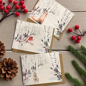 May include: Three winter-themed greeting cards with illustrations of a fox, a cardinal, and a rabbit in a snowy forest. The cards have a white background with birch trees and are accented with red berries and greenery. The cards are on brown cardstock.