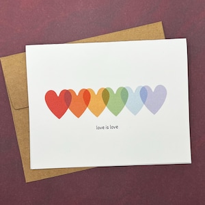 May include: A white greeting card with a row of overlapping hearts in rainbow colors: red, orange, yellow, green, blue, and purple. The words "love is love" are printed below the hearts. The card is inside a brown envelope.