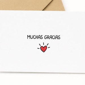 Pode incluir: Um cartão de felicitações retangular branco com o texto preto MUCHAS GRACIAS centralizado na frente. Abaixo do texto, há uma pequena ilustração de um coração vermelho com linhas pretas radiantes. O cartão está posicionado em frente a um envelope bege.