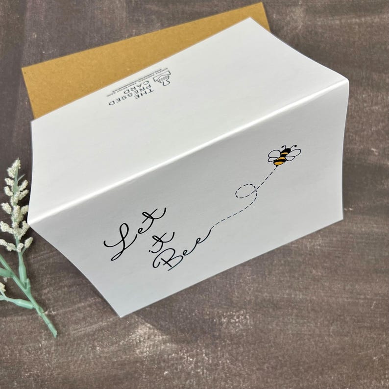 May include: White greeting card with the text "Let it Bee" in black script. A cartoon bee flies along a dotted line. The card is in a white box with a brown envelope.