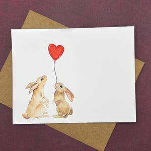 May include: A white greeting card featuring a watercolor illustration of two light brown rabbits standing on their hind legs and looking up at a single red heart-shaped balloon. The card is placed on top of a brown paper envelope.