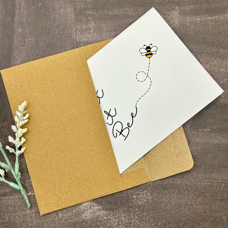 May include: A white greeting card featuring a cartoon bee and the words "Bee It" in black script, inside a brown envelope. A sprig of white flowers is in the lower left corner. The card is on a dark brown surface.