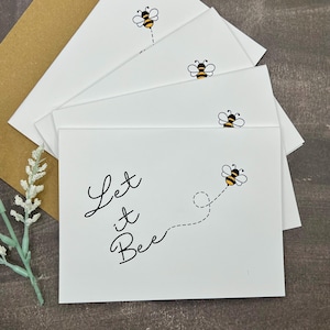 May include: White greeting cards featuring a bee illustration and the words "Let it Bee" in black script. The cards are arranged in a stack, with a brown envelope visible. A sprig of white flowers is in the lower left corner.