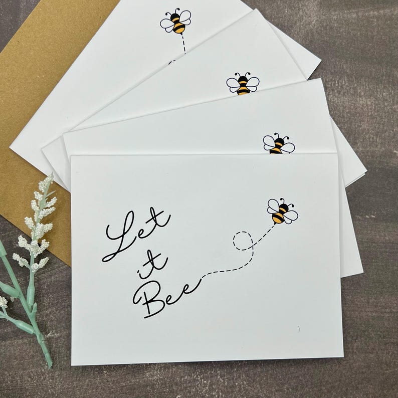 May include: White greeting cards featuring a bee illustration and the words "Let it Bee" in black script. The cards are arranged in a stack, with a brown envelope visible. A sprig of white flowers is in the lower left corner.