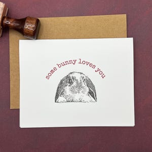 Può includere: Un biglietto di auguri bianco con la scritta "some bunny loves you" in rosso sopra un'illustrazione in bianco e nero di un coniglietto. Il biglietto è su una busta marrone, con un timbro di legno nell'angolo in alto a sinistra.