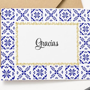 May include: A rectangular white greeting card featuring a blue and white floral tile pattern border and a gold decorative frame surrounding the word Gracias written in a black script font, accompanied by a brown paper envelope.