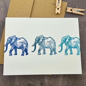 Puede incluir: Tarjeta de felicitación blanca con tres ilustraciones de elefantes azules. Los elefantes están representados en diferentes tonos de azul. La tarjeta está acompañada de un sobre marrón y pinzas de madera.