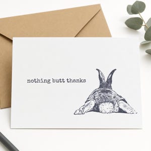 Pode incluir: Um cartão de felicitações branco com uma ilustração a tinta cinzento-escura de um coelho visto de trás, mostrando as orelhas, as patas traseiras e a cauda felpuda. O texto "nothing butt thanks" está impresso à esquerda do coelho.