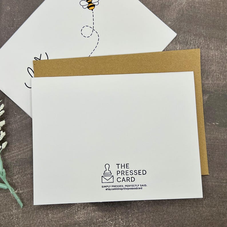 May include: A white greeting card featuring a bee illustration and the word "love" in black script. The card is layered over a brown envelope and a second white card. The back of the card has the text "THE PRESSED CARD".