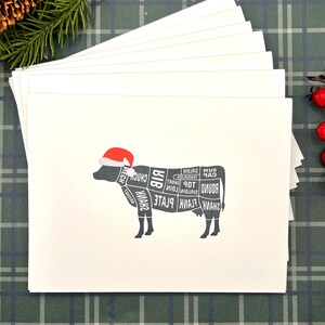 May include: A stack of white greeting cards with a black cow illustration wearing a red Santa hat. The cow graphic displays various meat cuts. The cards are on a green and white plaid surface, with pine and red berries in the background.