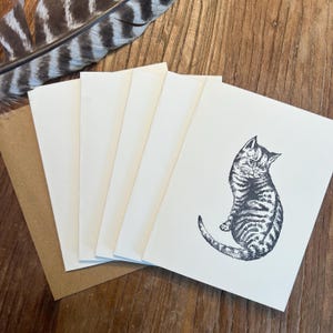 Peut inclure: Un ensemble de cartes de vœux vierges avec une illustration au stylo et encre d'un chaton tabby. Les cartes sont blanches avec une enveloppe marron et une plume sur une surface en bois.