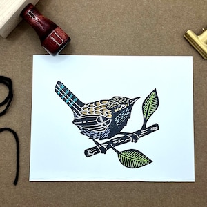 May include: A white card featuring a black and yellow linocut print of a bird perched on a branch with green leaves. A wooden stamp and a brass clip are visible in the background. Black string is in the lower left corner.