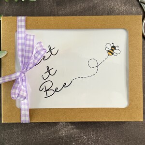May include: A brown rectangular box with a white interior, featuring the phrase "Let it Bee" and a cartoon bee. A light purple gingham ribbon is tied around the box. Scissors and greenery are in the background.