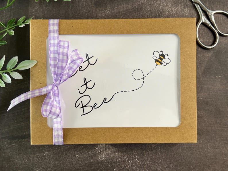 May include: A brown rectangular box with a white interior, featuring the phrase "Let it Bee" and a cartoon bee. A light purple gingham ribbon is tied around the box. Scissors and greenery are in the background.