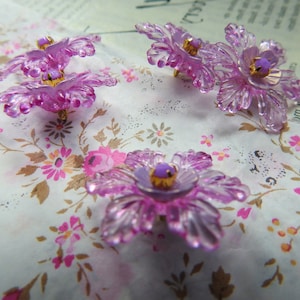 May include: Four purple plastic flower embellishments with gold centres. The flowers are on a white background with a floral pattern.