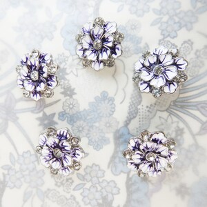 May include: Five flower-shaped decorative buttons. Each button has a white and purple enamel design with a silver-coloured metal rim and clear crystal accents. The buttons are arranged on a floral patterned surface.