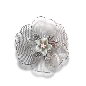 May include: A silver flower brooch with an intricate, openwork design. The brooch has a large flower shape with multiple petals and a smaller, central flower in muted green with a pink centre. The brooch is set against a white background.
