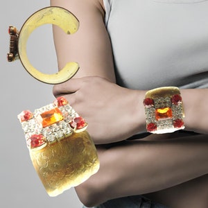 Può includere: Due braccialetti dorati con accenti di pietre preziose rosse e arancioni. I braccialetti sono entrambi decorati e presentano un design quadrato con una grande pietra centrale.