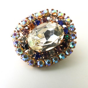 May include: A vintage brooch featuring a large, clear, faceted crystal at its centre, encircled by a halo of smaller, colourful rhinestones. The rhinestones are in shades of blue, purple, orange, and green, set in a gold-tone metal frame.