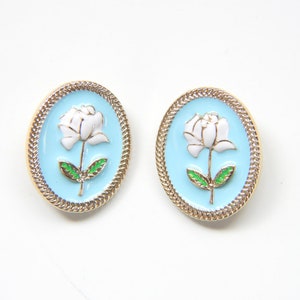 May include: A pair of gold-tone earrings with a white flower design on a light blue background. The earrings are oval shaped and have a gold rope border.