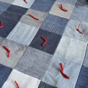 Puede incluir: Una colcha de retazos hecha de cuadrados de mezclilla con detalles de hilo rojo. La colcha está hecha de diferentes tonos de mezclilla azul.