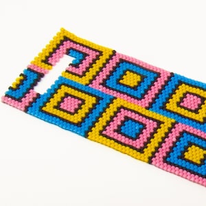 May include: A colourful beaded bracelet with a geometric pattern of pink, blue, yellow and black squares. The bracelet has a clasp closure.