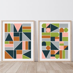 May include: Two framed prints with geometric abstract designs in shades of green, pink, brown, and blue. The prints are hung on a white wall.