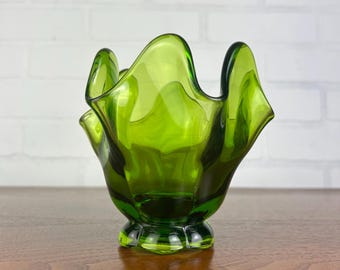 May include: A vibrant green glass bowl with a unique, scalloped rim. The bowl has a rounded base and a flared, petal-like design. The glass is a rich, translucent green, and the bowl sits on a wooden surface against a white brick wall.